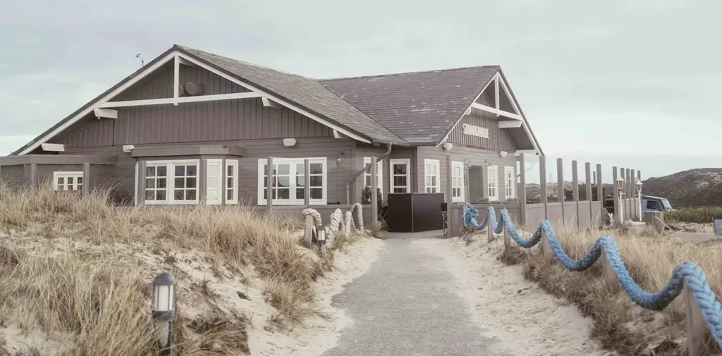 Außenansicht der Strandhalle Sylt mit dunkler Holzfassade, Dünenlandschaft und Weg zum Restaurant direkt an der Nordsee.