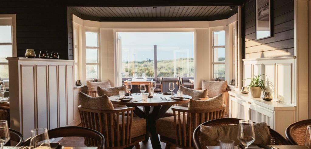 Eleganter Restaurantbereich der Strandhalle Sylt mit rundem Holztisch, gepolsterten Sesseln und Blick auf die Dünenlandschaft von Sylt.