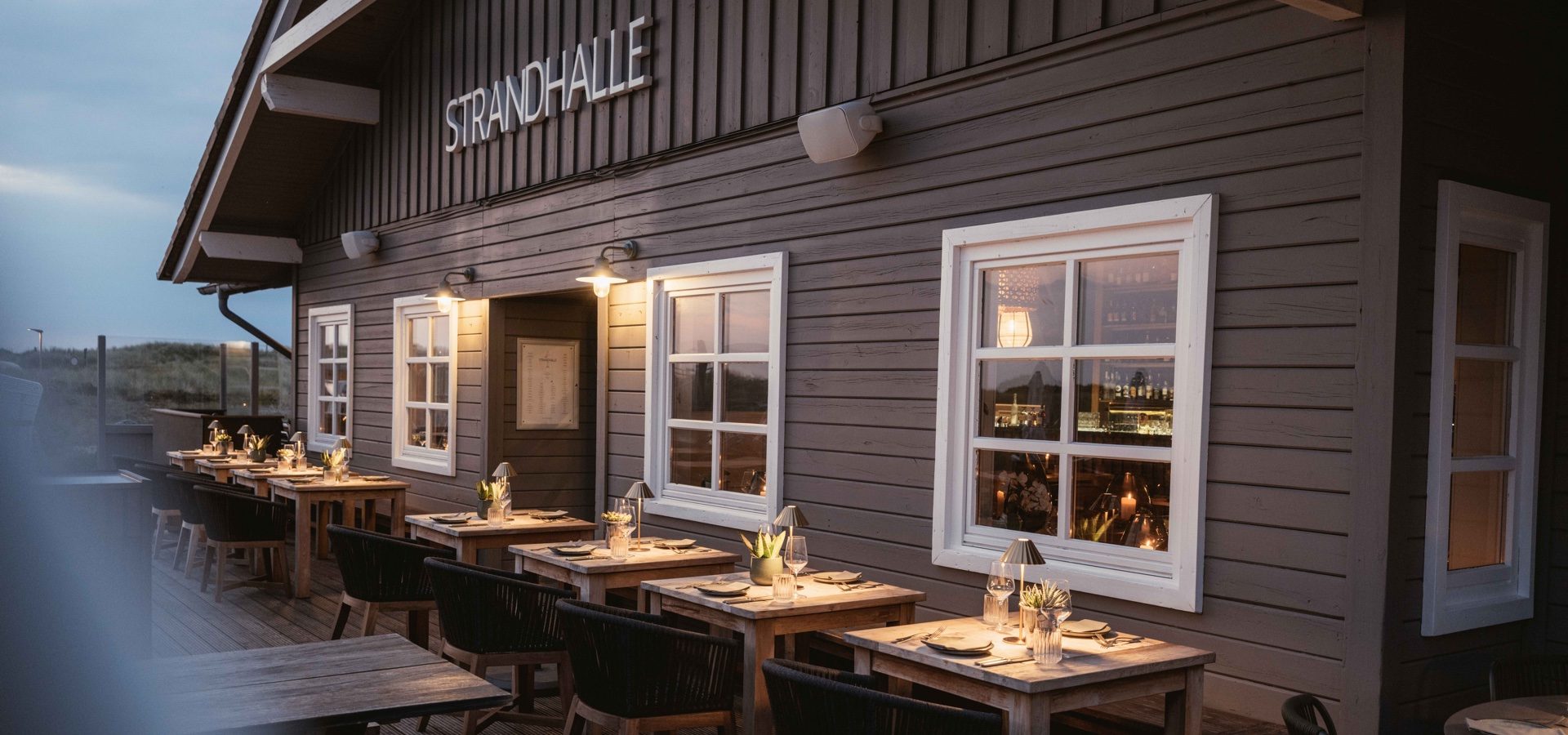 Terrasse der Strandhalle Sylt in warmer Abendstimmung mit Holztischen, Außenbeleuchtung und Blick auf die Dünenlandschaft.